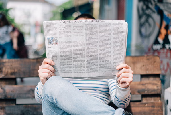 Photo of person seated on an outdoor bench reading a newspaper