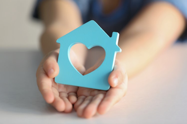 Photo of a child's hands holding a house-shaped, blue wooden block, with a heart cutout in the center