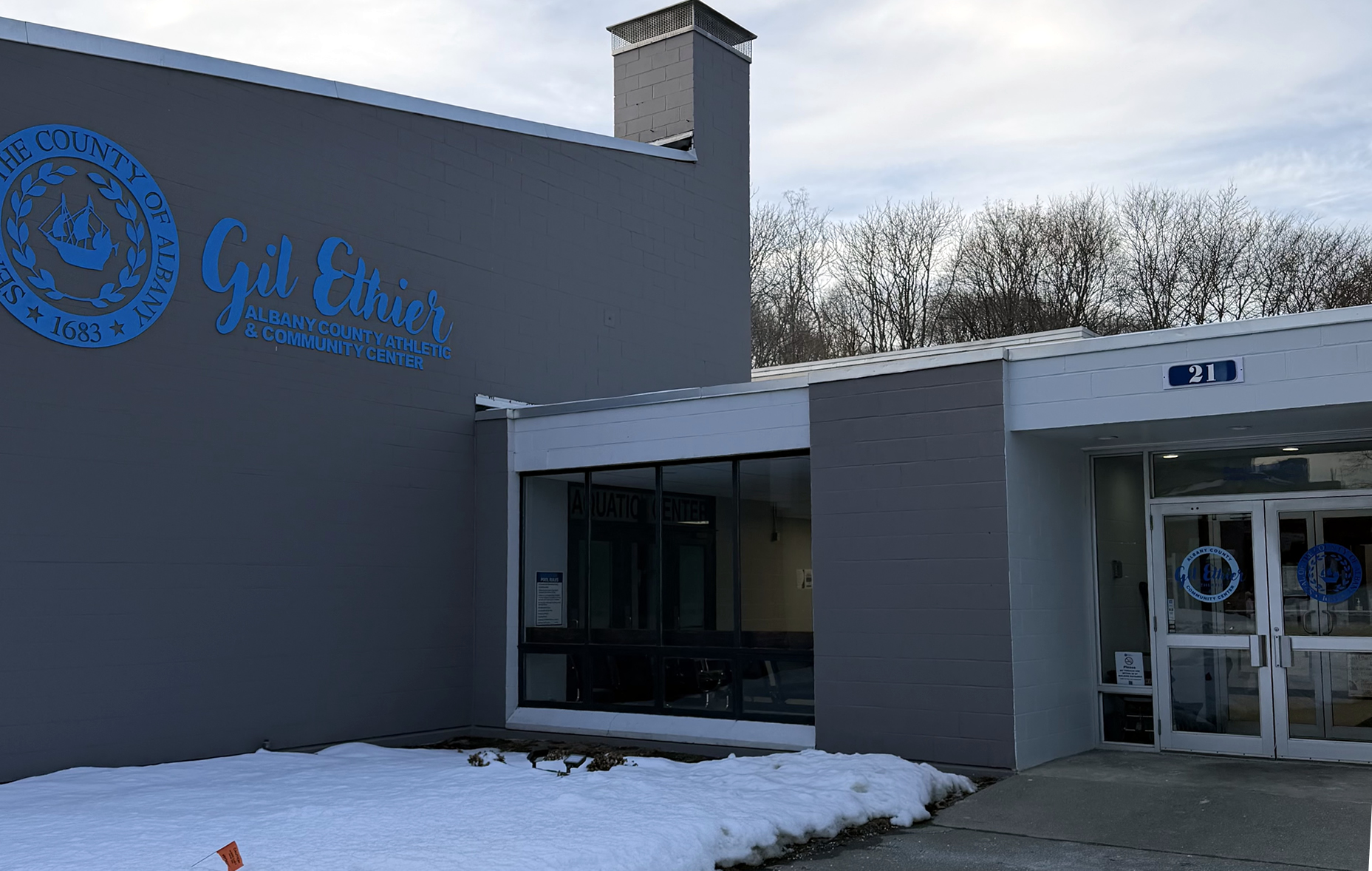 Photo of the outside of The Gil Ethier Community Center, showing the name on the side of the building and the front doors. There is snow on the ground.
