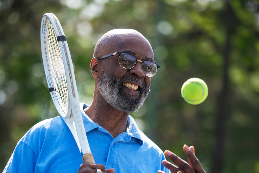 Photo of smiling man outdoors with tennis racket in one hand, tossing tennis ball upwards with other hand.